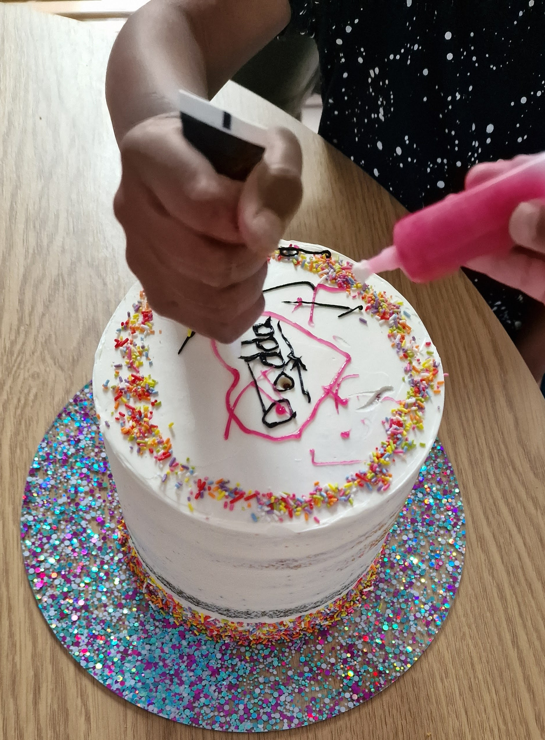 Hands-on process of adding pink and black icing writing and sprinkles to a fresh custom cake in our Greenford studio kitchen